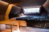 Experience comfort in this cozy RV rental interior, showcasing a compact bed, forest window view, and ambient lighting.