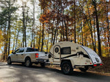 RV rental: A pickup truck tows a white teardrop camper on a sunny autumn road through a vibrant forest, ideal for camping adventures.