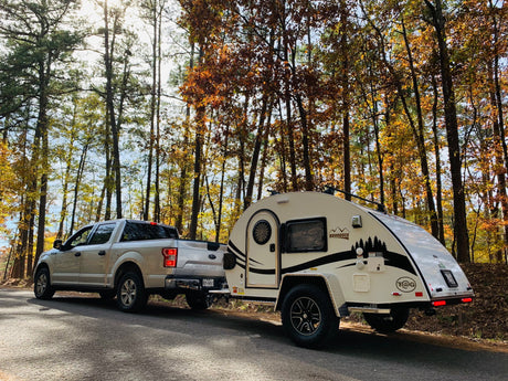 RV rental: A pickup truck tows a white teardrop camper on a sunny autumn road through a vibrant forest, ideal for camping adventures.