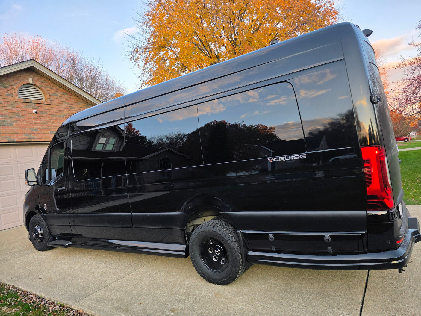 This sleek black VCRUISE RV rental van, customized for adventure, is parked on a driveway amidst autumn colors.