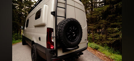 Modern campervan for rv rental, featuring a rear spare tire & ladder, parked by a forest road. Perfect for adventure.