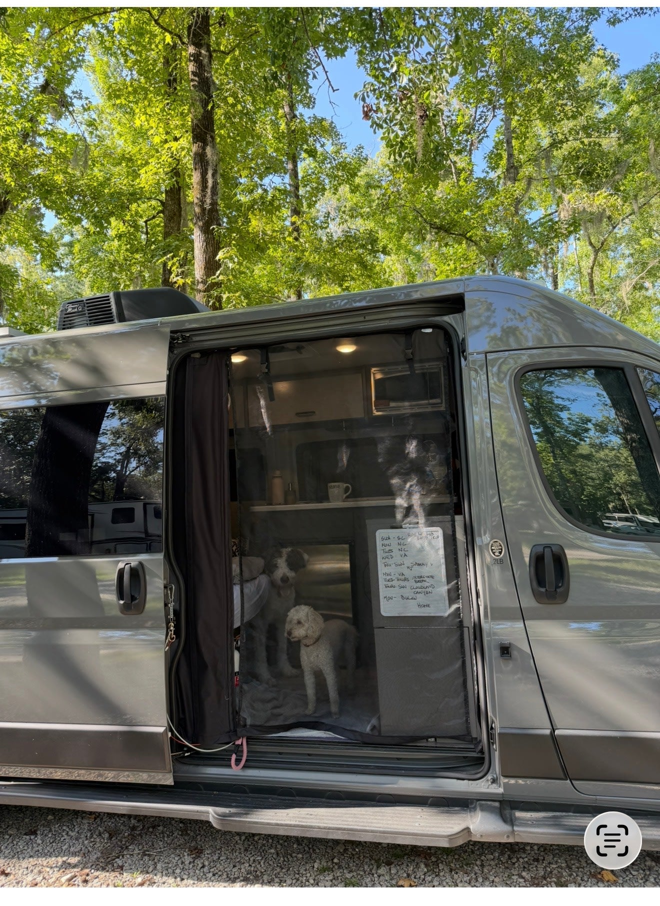 An RV rental camper van with an open screen door, revealing two dogs inside and a trip schedule, parked amongst trees.