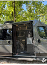 An RV rental camper van with an open screen door, revealing two dogs inside and a trip schedule, parked amongst trees.