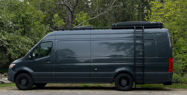 Explore with this dark gray Mercedes Sprinter RV rental. A custom camper van with roof rack & ladder, parked in nature.