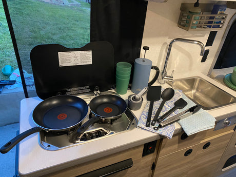 RV rental kitchen setup: non-stick pans on a stovetop, a sink, French press, cups, and cooking utensils ready for use.