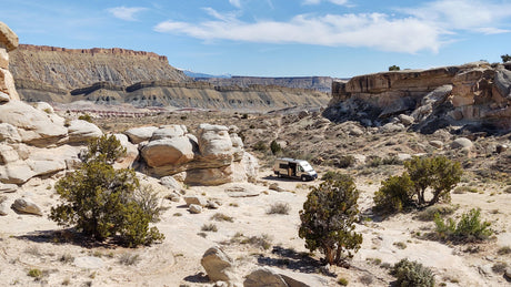 An RV rental is perfectly situated amidst unique desert rock formations, with layered mesas and distant snowy peaks under clear skies.