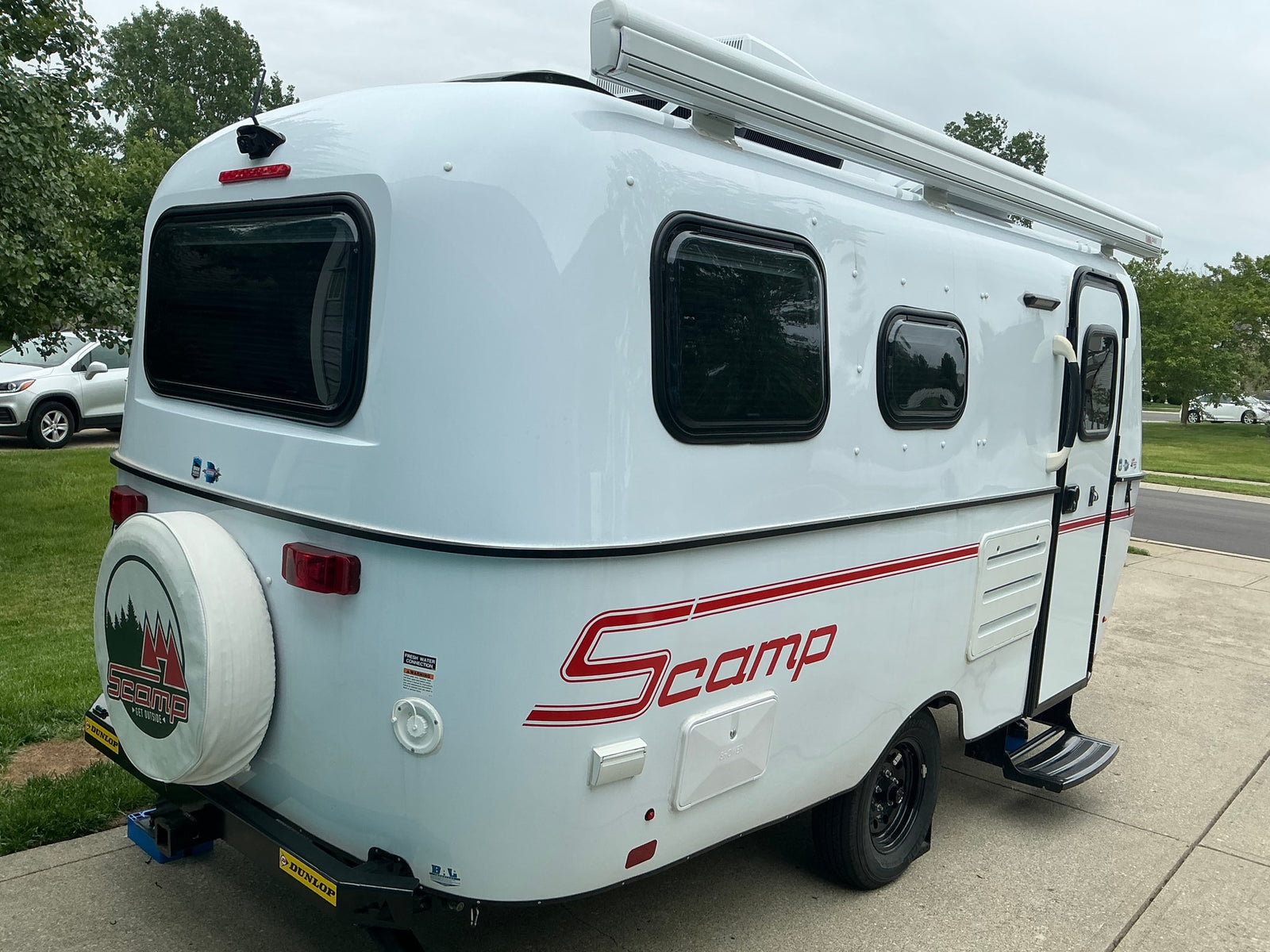 Rear-side view of a white Scamp travel trailer, featuring a spare tire, awning, and iconic red stripe. Ideal for an RV rental.