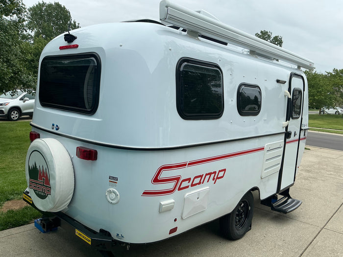Rear-side view of a white Scamp travel trailer, featuring a spare tire, awning, and iconic red stripe. Ideal for an RV rental.