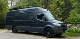 Dark gray Mercedes Sprinter camper van with roof rack & cargo box, ready for rv rental adventures, parked by a forest.