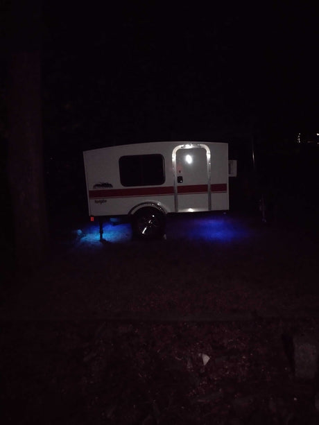 A white teardrop-style camper, an ideal rv rental, is illuminated by blue underglow and door lights at night, parked on dark ground.