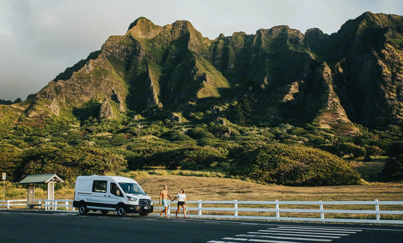 Exploring nature with an RV rental. A couple walks by their white camper van, with impressive green mountains in the background.