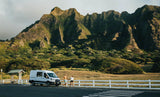 Exploring nature with an RV rental. A couple walks by their white camper van, with impressive green mountains in the background.