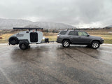 Grey SUV towing a light grey teardrop RV rental, ready for mountain adventures on a wet road.