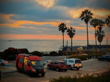 Red RV rental by the ocean at sunset, with palm trees and a train passing. A beautiful coastal scene.
