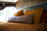 Inside an RV rental, a bed with golden and brown pillows, including a textured bolster, beneath wooden storage cabinets.