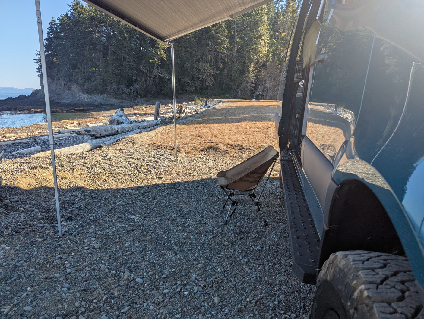 An RV rental parked at a scenic coastal campsite with an awning, chair, and driftwood on the rocky beach.