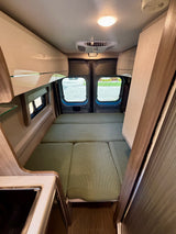 Cozy rv rental interior with a convertible bed, rear windows, overhead storage, and a glimpse of the kitchenette.