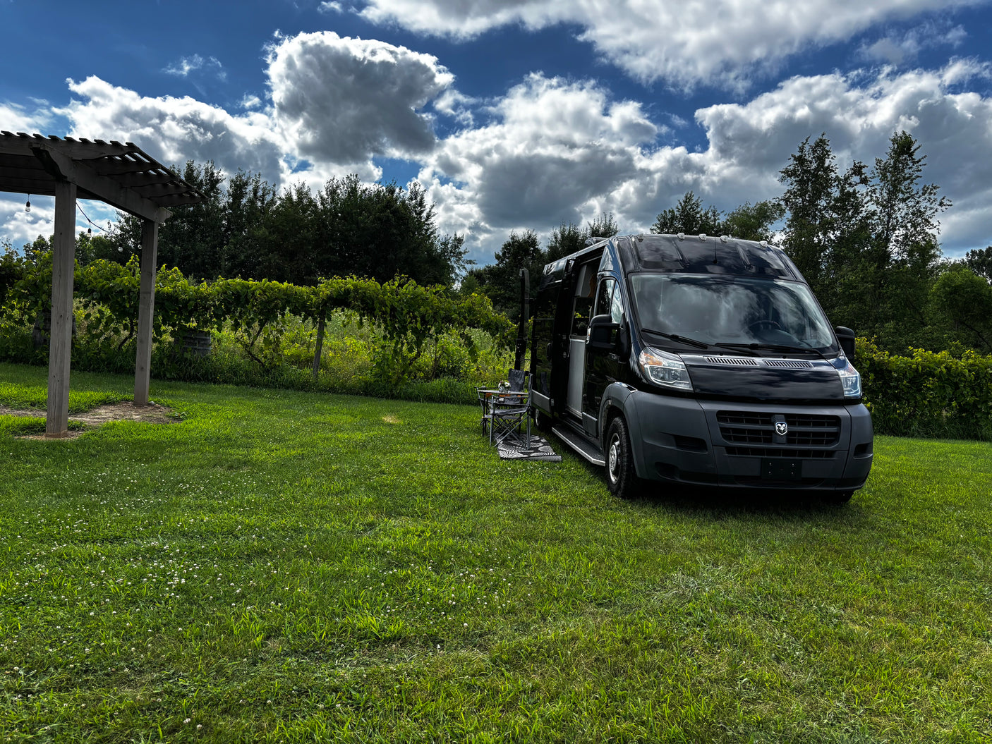 An RV rental camper van parked in a vibrant vineyard with a pergola and outdoor seating under a bright, cloudy sky.