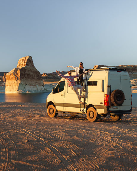 Happy couple posing on their rv rental camper van roof, overlooking a scenic lake and desert rock formations at sunset. Adventure awaits!