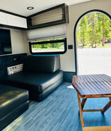 Modern RV rental interior with black seating, a folding wood table, and a bright view of the forest outside.