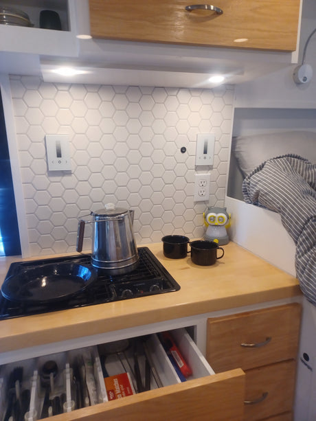 RV rental kitchen: compact stovetop, coffee pot, and organized open drawers with utensils. Smart controls on hex tile backsplash.