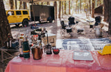 A perfect rv rental camp kitchen setup: red checkered table, coffee gear, and a yellow Jeep in a sun-dappled forest.