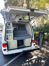 An **RV rental** camper van with its tailgate open, revealing a convertible bed, built-in storage, and a rear bike rack.