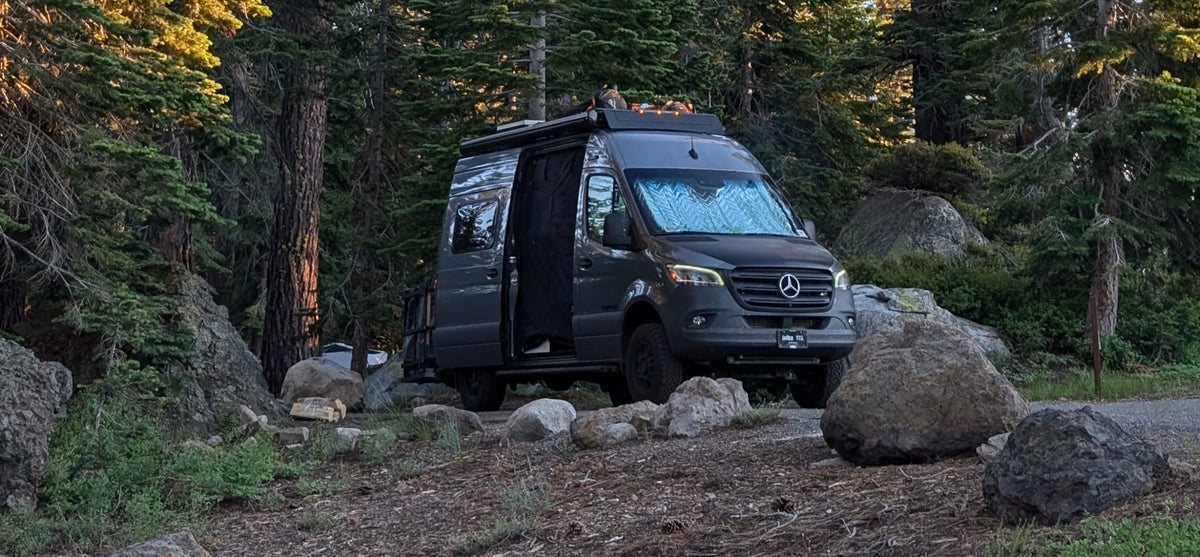 An rv rental Sprinter camper van parked among pine trees and large rocks at a serene campsite. Door open, sunshade visible. — photo 2