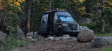 An rv rental Sprinter camper van parked among pine trees and large rocks at a serene campsite. Door open, sunshade visible.