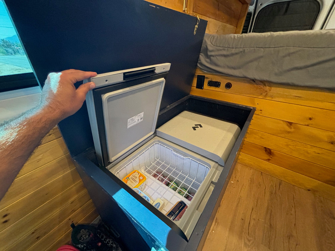 Inside a cozy **rv rental**: a hand opens a built-in chest fridge filled with groceries, next to a matching covered unit.