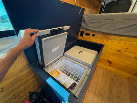 Inside a cozy **rv rental**: a hand opens a built-in chest fridge filled with groceries, next to a matching covered unit.