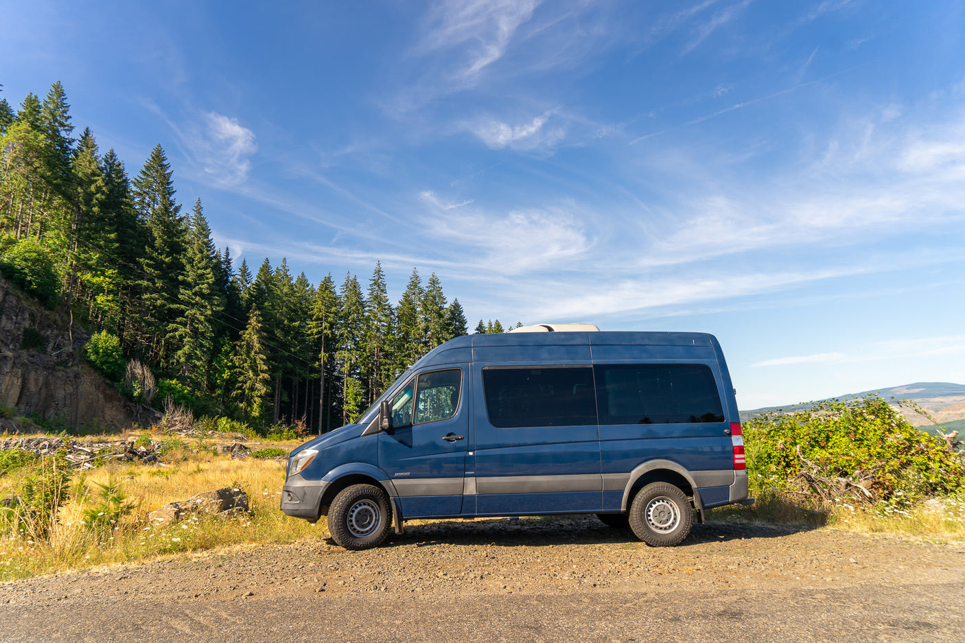 Blue Sprinter van, likely an RV rental, parked on a scenic dirt road with tall trees and a blue sky. Perfect for a road trip!