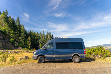 Blue Sprinter van, likely an RV rental, parked on a scenic dirt road with tall trees and a blue sky. Perfect for a road trip!
