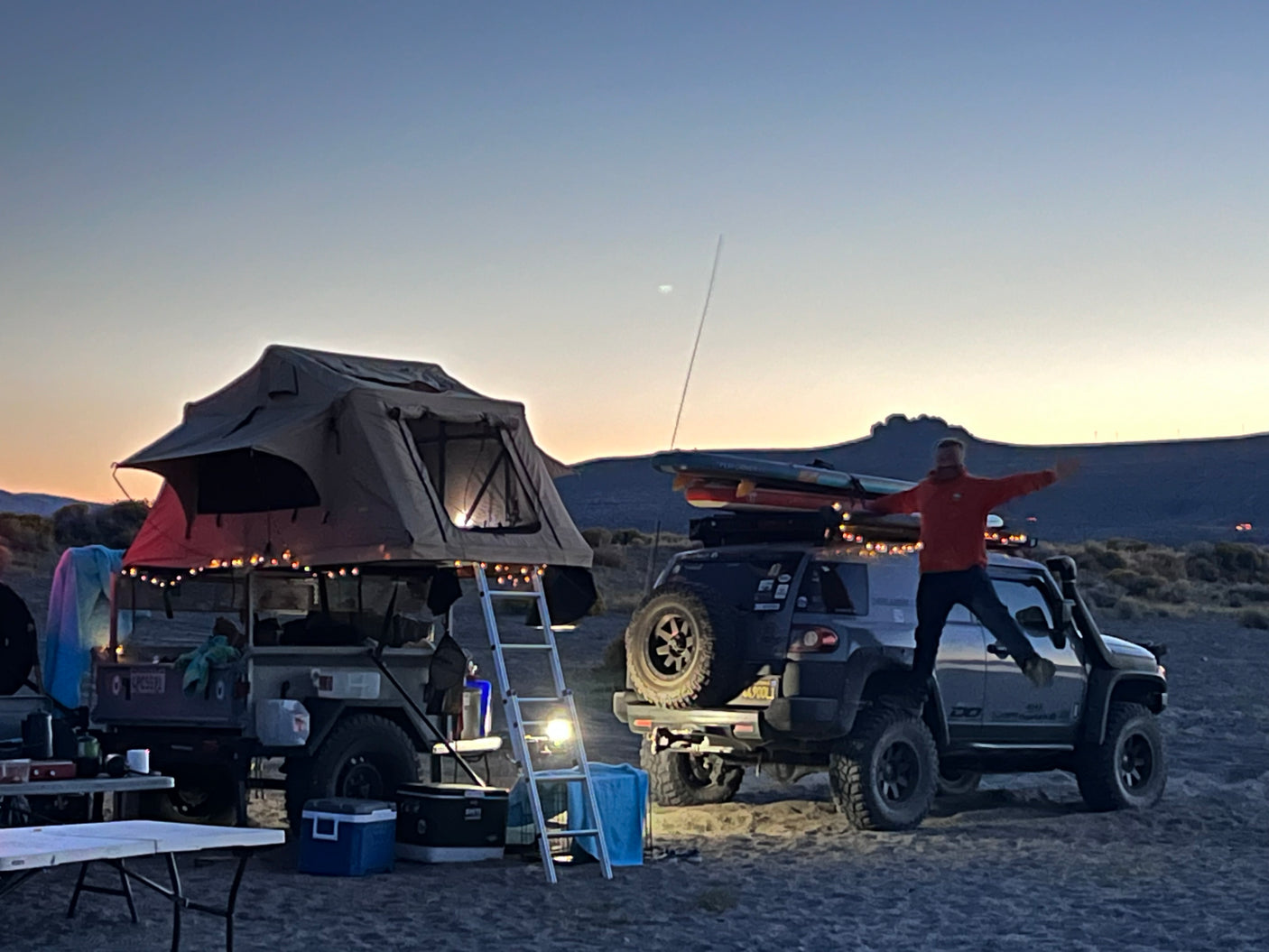 Dusk camping: A person jumps on an SUV next to a trailer with a lit rooftop tent. This setup offers adventures similar to an RV rental.