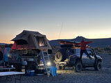 Dusk camping: A person jumps on an SUV next to a trailer with a lit rooftop tent. This setup offers adventures similar to an RV rental.