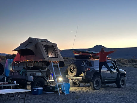 Dusk camping: A person jumps on an SUV next to a trailer with a lit rooftop tent. This setup offers adventures similar to an RV rental.