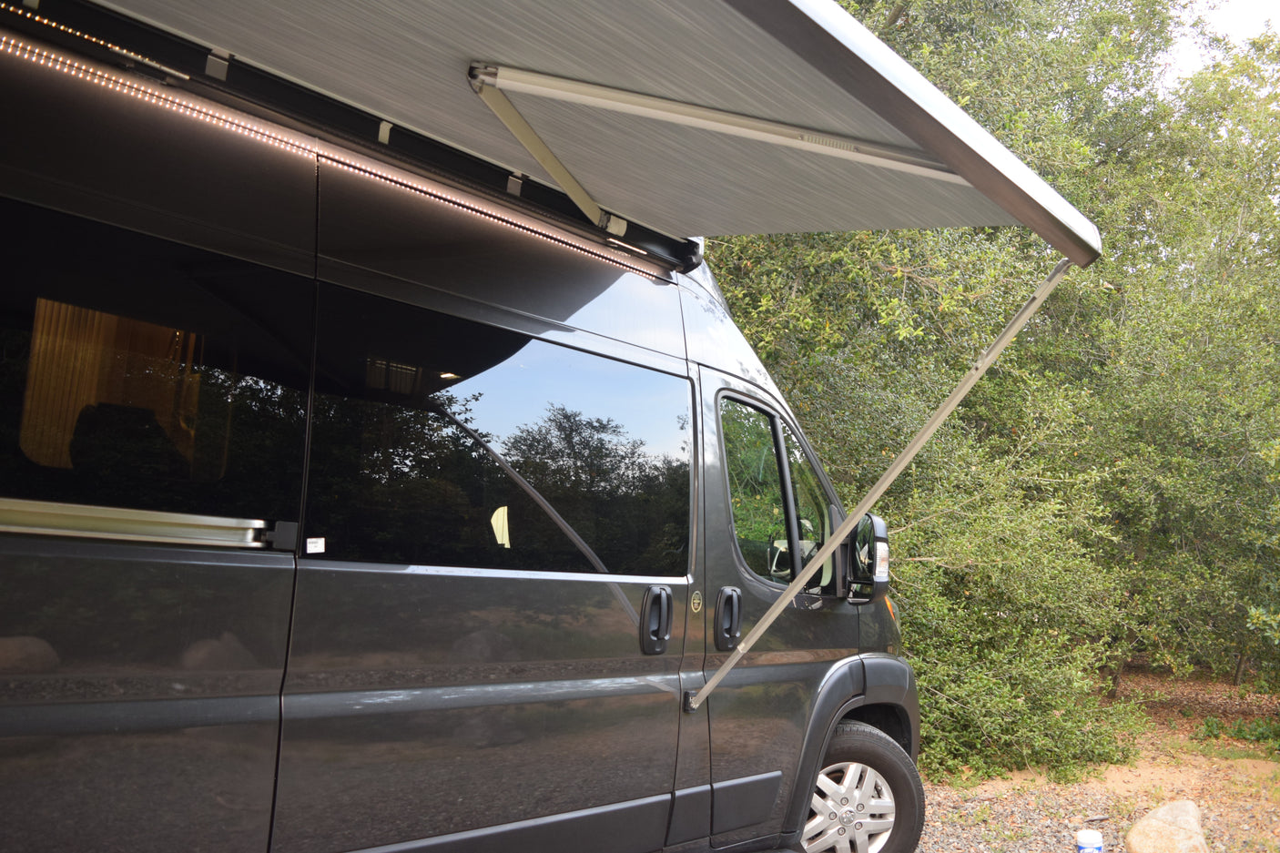 Side view of a grey RV rental with its awning extended and bright LED lights on, parked amidst green trees.