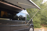 Side view of a grey RV rental with its awning extended and bright LED lights on, parked amidst green trees.