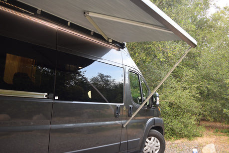 Side view of a grey RV rental with its awning extended and bright LED lights on, parked amidst green trees.