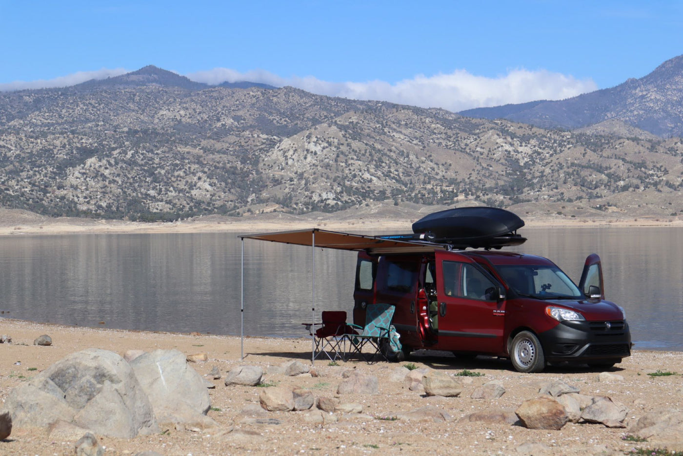 Red RV rental van camping by a lake with mountains, awning extended for outdoor relaxation. Perfect for van life adventures!