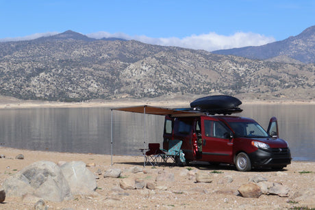 Red RV rental van camping by a lake with mountains, awning extended for outdoor relaxation. Perfect for van life adventures!