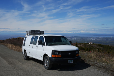 White RV rental van with rooftop tent on a scenic Alaska mountain road, overlooking the city and mountains.
