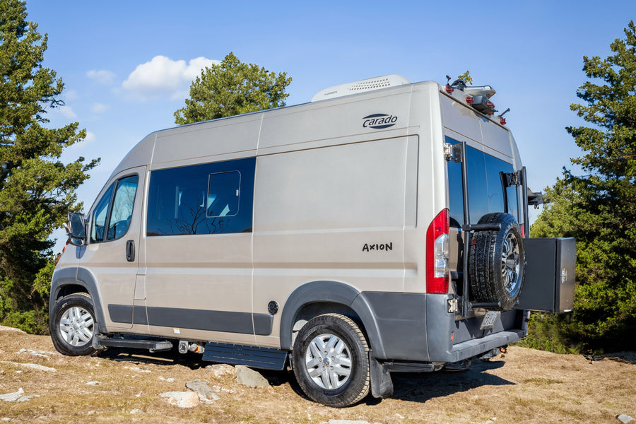 A tan Carado Axion camper van, equipped with a spare tire and roof AC, perfect as an RV rental for outdoor adventures.