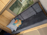 RV rental: Cozy camper van interior with a gray bench seat, seatbelts, and decorative pillows by light wood cabinets and a large window.
