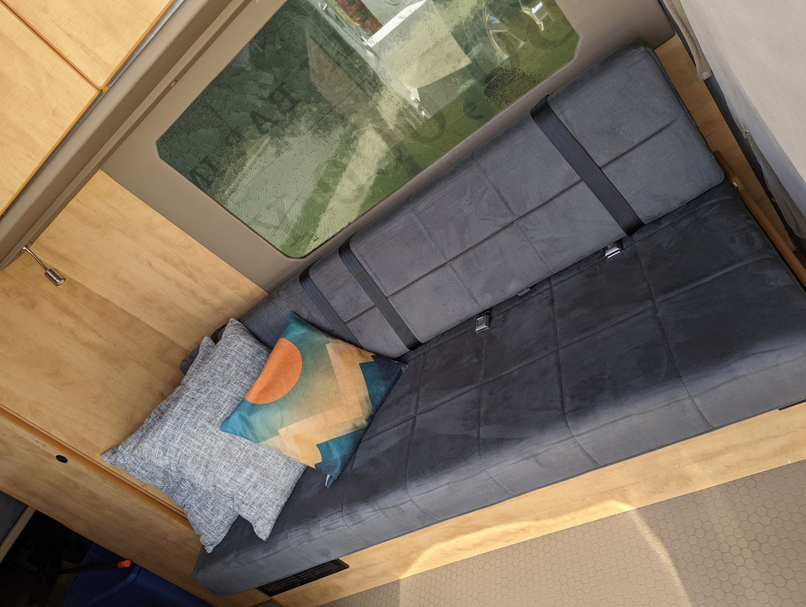 RV rental: Cozy camper van interior with a gray bench seat, seatbelts, and decorative pillows by light wood cabinets and a large window.