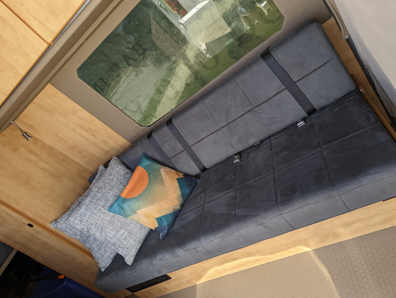 RV rental: Cozy camper van interior with a gray bench seat, seatbelts, and decorative pillows by light wood cabinets and a large window. — photo 6