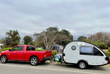 A red pickup truck tows a gray teardrop RV rental camper at a campsite under an overcast sky.