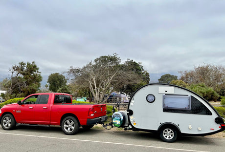 A red pickup truck tows a gray teardrop RV rental camper at a campsite under an overcast sky.