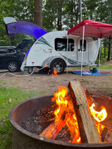 A warm campfire glows, perfectly complementing this cozy teardrop RV rental setup at a wooded campground.