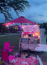 Cozy pink-themed teardrop RV rental setup at dusk, featuring string lights, camping chairs, and a flamingo rug.